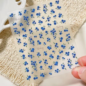 May include: A transparent sheet of nail art stickers featuring a repeating pattern of small blue blueberries with green leaves. The word Blueberry is printed in a cursive script in three locations on the sheet, and the code YT8601 is in the corner.