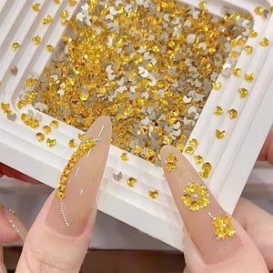 Könnte beinhalten: Nahaufnahme eines weißen Behälters gefüllt mit goldenen und silbernen Nagelkunst-Strasssteinen. Zwei lange, manikürte Nägel sind mit goldenen Strasssteinen in verschiedenen Designs verziert, darunter eine Linie und Blumenmuster.