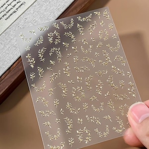 May include: A rectangular, translucent sheet featuring numerous small, metallic gold adhesive decals in various botanical leaf and vine patterns. The stickers are arranged in a grid-like formation on the clear, frosted backing material.