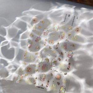 Puede incluir: Lámina transparente de pegatinas para uñas con diseños de conchas marinas y estrellas en colores dorados, rosas e iridiscentes. Las pegatinas incluyen el texto "Alice" y "YT5871Pro". La lámina está sobre una superficie blanca.