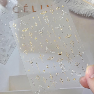 May include: Clear nail art stickers featuring gold and silver celestial designs. The sheet includes stars, moons, crosses, and decorative elements. The text "YT7101Me" is printed on the bottom left corner of the sheet.