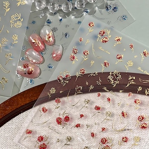 May include: Clear nail art stickers featuring gold and red rose designs. Some stickers have a gold outline, while others are filled with red. Several nail designs with rose stickers are also visible.