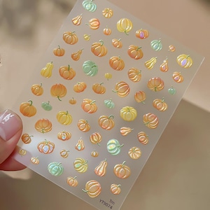 May include: A rectangular sheet of adhesive nail decals featuring a variety of small pumpkins and gourds in shades of orange, yellow, and light green. The designs have a glossy, iridescent finish. The text YT9074 Tm is printed in the bottom corner.