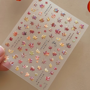 May include: A rectangular sheet of adhesive nail decals featuring numerous small, iridescent maple leaf designs in shades of red, orange, yellow, and gold. The sheet includes various Chinese characters and the text "Te YT9213" printed in black.