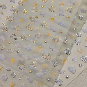 以下が含まれることがあります： 金色の縁取りの雲、太陽、星が繰り返されるパターンのネイルアートステッカー。雲は白とラベンダー色です。「Me YT7790」の文字が1枚に印刷されています。ステッカーは透明な台紙に貼られています。