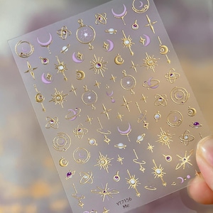 Puede incluir: Lámina transparente de pegatinas para uñas con diseños celestiales dorados. Las pegatinas incluyen lunas, estrellas, planetas y soles con detalles morados y transparentes. El texto "YT7156 Me" está impreso en la esquina inferior derecha.