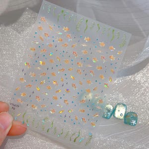 May include: Clear nail art sticker sheet featuring small orange fish, blue accents, and green seaweed designs. The sheet includes the text "YT5079 X9". The stickers are shown next to painted nails with a similar aquatic theme.