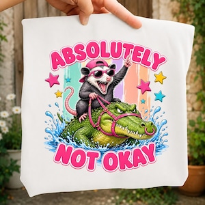 Può includere: Una borsa tote bianca con la grafica di un opossum che indossa un berretto rosa e occhiali da sole, a cavallo di un alligatore verde con una pettorina rosa. Il testo ABSOLUTELY NOT OKAY è scritto in rosa, circondato da stelle e spruzzi d'acqua.