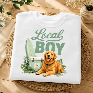 May include: A white crewneck sweatshirt featuring a graphic print of a golden retriever lying down next to a light green cooler and a surfboard. The text "Local BOY" is printed in a sage green font above the dog, with palm leaves surrounding the base.