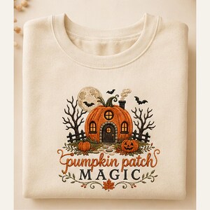 Op de afbeelding: Een crèmekleurig sweatshirt met een borduursel van een pompoenvormig huis met ramen en een deur. De afbeelding bevat een volle maan, kale bomen, vleermuizen en kleine pompoenen. De tekst luidt "pumpkin patch MAGIC" in oranje en zwarte letters.