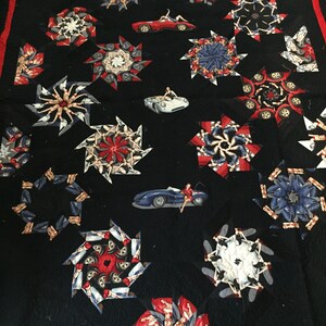 May include: Black quilt with red trim featuring repeating starburst patterns and car motifs. The starbursts are composed of images of women and cars in red, white, and blue. Several car images are scattered across the quilt.