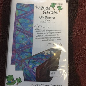 May include: A clear plastic package containing a sewing pattern for an Obi Runner. The pattern is labeled "Pagoda Garden Obi Runner" and measures 110" x 14". The pattern features a colorful geometric design with blue, purple, and green hues.