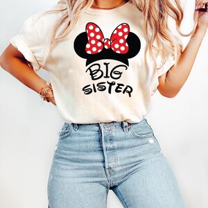 May include: Cream-colored t-shirt featuring a Minnie Mouse design with a red and white polka dot bow and the words "Big Sister" in black. The shirt is paired with light blue jeans.