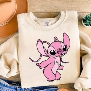May include: Cream-coloured sweatshirt with a pink cartoon character. The character has large ears, black eyes, and a white lightning bolt design. The sweatshirt is made of a soft material. The cartoon character has a playful expression.