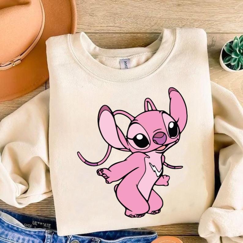 May include: Cream-coloured sweatshirt with a pink cartoon character. The character has large ears, black eyes, and a white lightning bolt design. The sweatshirt is made of a soft material. The cartoon character has a playful expression.