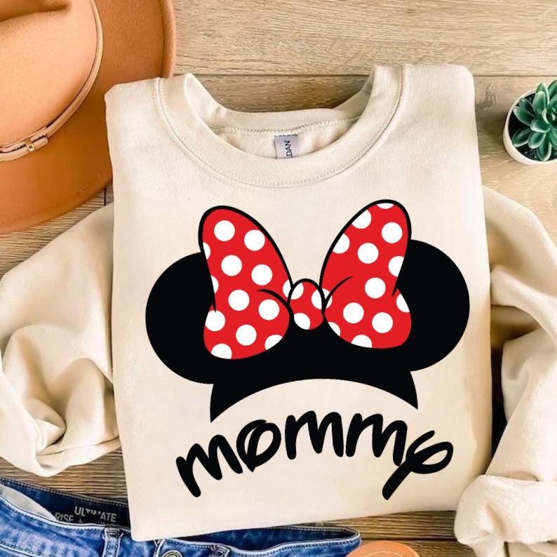 May include: Cream-colored sweatshirt featuring a black Minnie Mouse silhouette with a red polka-dot bow. The word "mommy" is written in a black script font below the graphic. The sweatshirt is a casual, comfortable style.