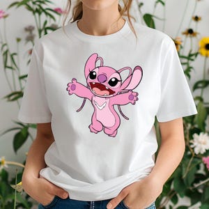 May include: White t-shirt with a pink cartoon character design. The character has large eyes, open arms, and a wide smile. The character has large ears and a light pink color scheme. The shirt is made of a soft material.