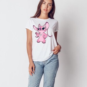 May include: White t-shirt with a pink cartoon character wearing sunglasses. The character has large ears and is in a pose with one hand on its hip. The t-shirt is worn with blue jeans.