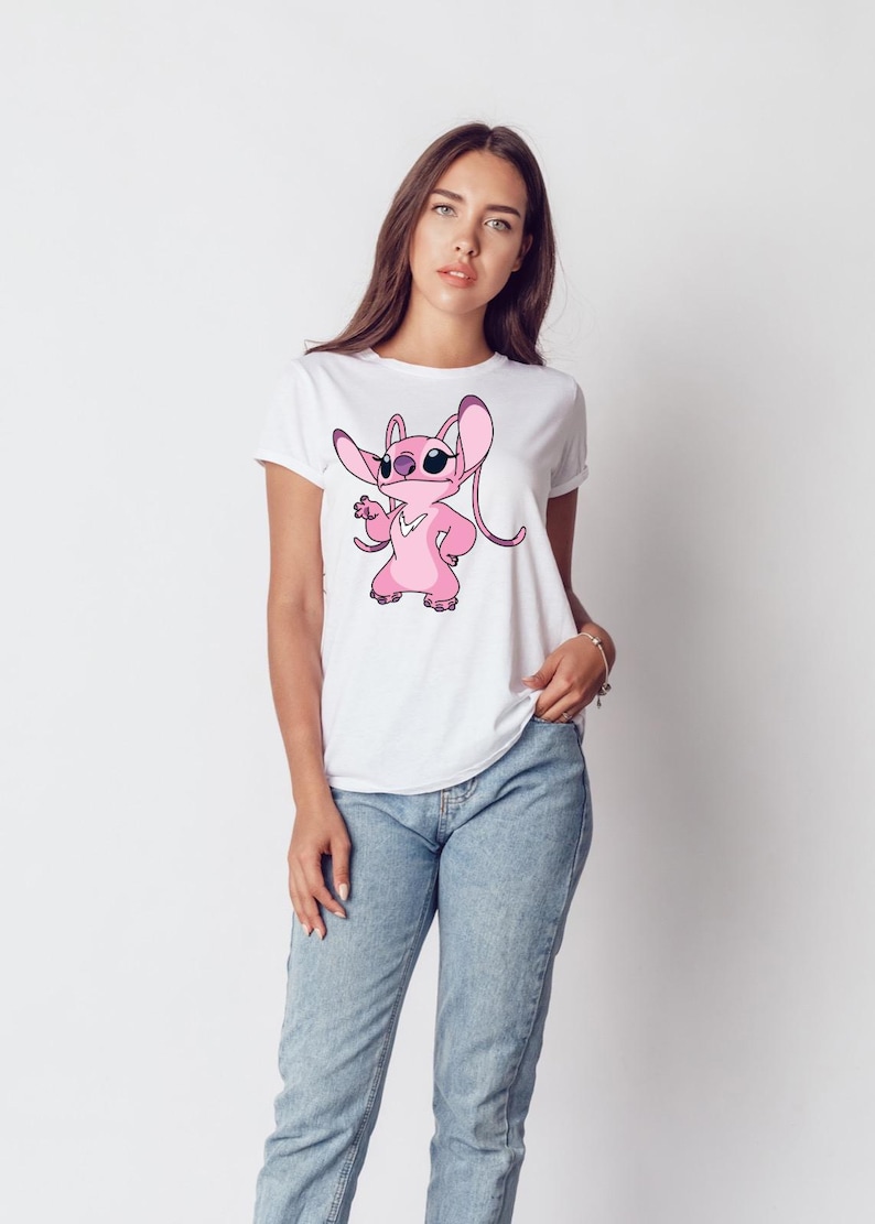 May include: White t-shirt with a pink cartoon character wearing sunglasses. The character has large ears and is in a pose with one hand on its hip. The t-shirt is worn with blue jeans.