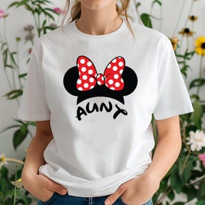 May include: White t-shirt featuring a black Minnie Mouse silhouette with a red polka dot bow. The word "AUNT" is printed in black below the silhouette. A fun and playful design.