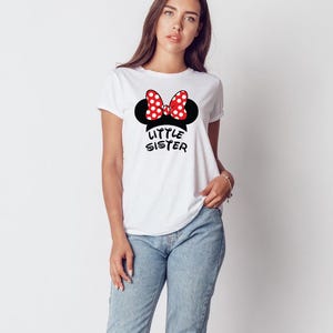 May include: White t-shirt with a black Minnie Mouse silhouette, a red and white polka dot bow, and the words "Little Sister" in black. The shirt is paired with blue jeans.
