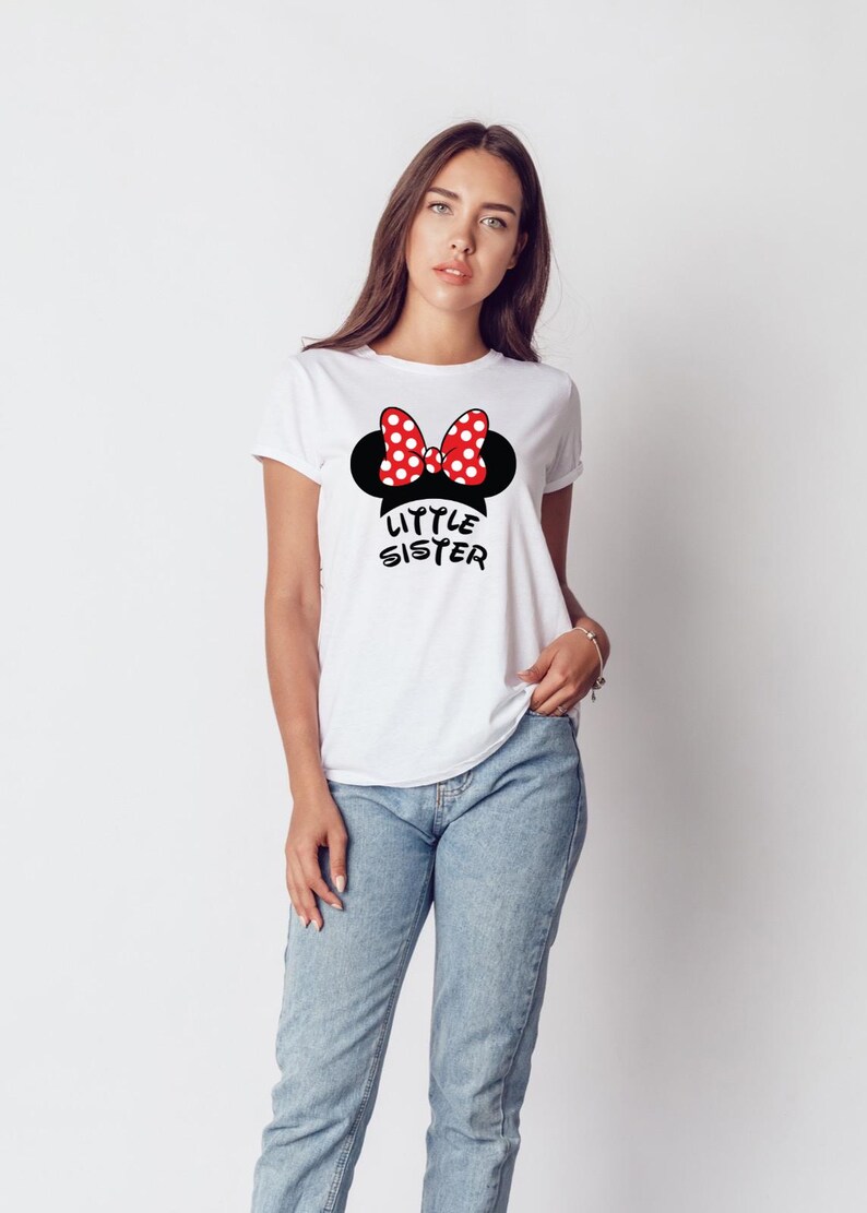 May include: White t-shirt with a black Minnie Mouse silhouette, a red and white polka dot bow, and the words "Little Sister" in black. The shirt is paired with blue jeans.