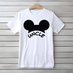 May include: White t-shirt with a black graphic of Mickey Mouse ears and the word "UNCLE" in a curved font. The shirt is hanging on a wooden hanger against a white and gray wood-paneled wall.
