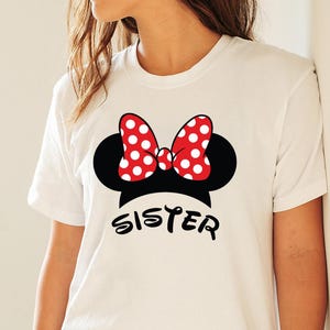 May include: White t-shirt featuring a black Minnie Mouse silhouette with a red polka-dot bow. The word "SISTER" is printed in black script below the design. The shirt is a crew neck and has short sleeves.