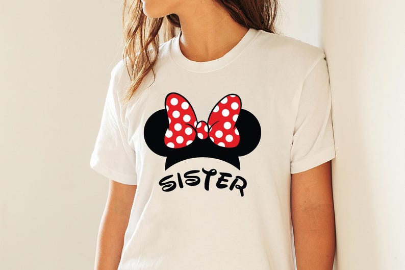 May include: White t-shirt featuring a black Minnie Mouse silhouette with a red polka-dot bow. The word "SISTER" is printed in black script below the design. The shirt is a crew neck and has short sleeves.