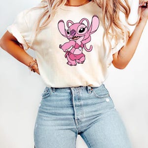May include: A cream-coloured t-shirt with a pink cartoon character wearing a pink bikini. The character has large ears and a playful expression. The t-shirt is paired with light blue denim jeans.