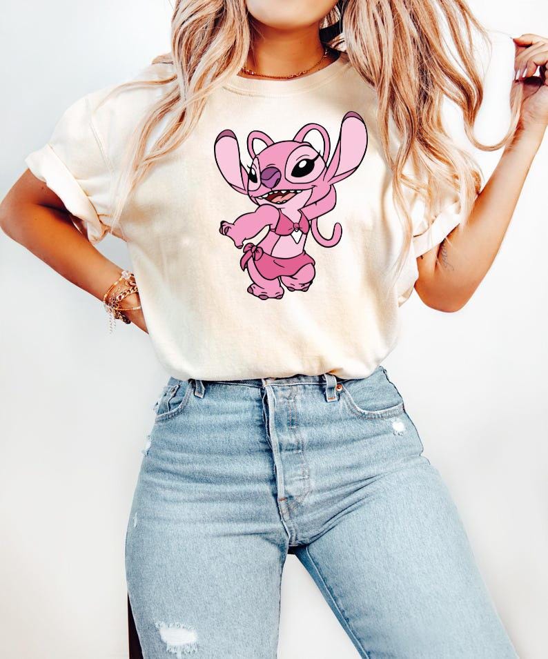 May include: A cream-coloured t-shirt with a pink cartoon character wearing a pink bikini. The character has large ears and a playful expression. The t-shirt is paired with light blue denim jeans.