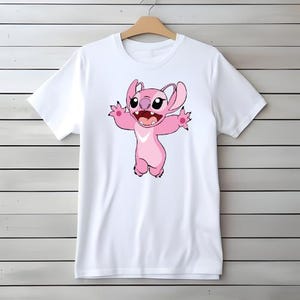 May include: White t-shirt with a pink cartoon character design. The character has large eyes, a wide smile, and outstretched arms. A white chevron is on the chest. The t-shirt is displayed on a wooden hanger against a light wood-panelled wall.