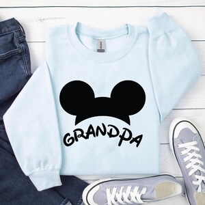 May include: A light blue sweatshirt with a black Mickey Mouse silhouette and the word "Grandpa" in a playful font. The sweatshirt is paired with blue jeans and gray sneakers on a white wooden surface.
