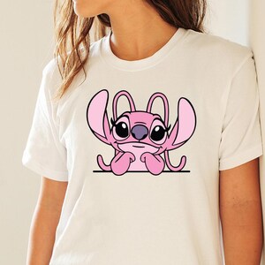 May include: White t-shirt with a pink cartoon character design. The character has large ears, black eyes, and a purple nose, resting its chin on its hands. The design is centered on the shirt.