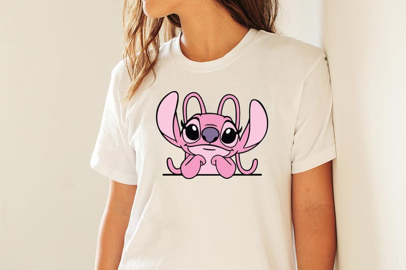 May include: White t-shirt with a pink cartoon character design. The character has large ears, black eyes, and a purple nose, resting its chin on its hands. The design is centered on the shirt.