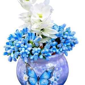 May include: A decorative floral arrangement in a round, lavender-coloured vase. The vase features a blue butterfly and white flowers. The arrangement includes white and blue flowers, creating a vibrant display. The image is set against a white background.