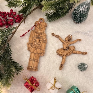 Może przedstawiać: Drewniane ozdoby świąteczne przedstawiające żołnierza-dziadka do orzechów i baletnicę. Ozdoby są jasnobrązowe z misternymi detalami i małymi otworami do zawieszania. Obecne są również pudełka na prezenty i zieleń.