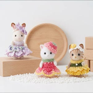 Pode incluir: Tr&ecirc;s pequenas figuras de animais vestidas com roupas de croch&ecirc; feitas &agrave; m&atilde;o. Um coelho usa um vestido de babados lavanda com acess&oacute;rio de cabe&ccedil;a, um gato branco usa um vestido rosa e verde com chap&eacute;u, e um gato malhado usa um vestido amarelo e verde.