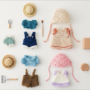 Pode incluir: Uma cole&ccedil;&atilde;o de roupas e acess&oacute;rios de croch&ecirc; em miniatura para bonecas, incluindo dois chap&eacute;us de palha, duas toucas em creme e rosa, v&aacute;rios tops, jardineiras e shorts em azul, marrom e branco, al&eacute;m de uma pequena p&aacute;, um ancinho e blocos de madeira.