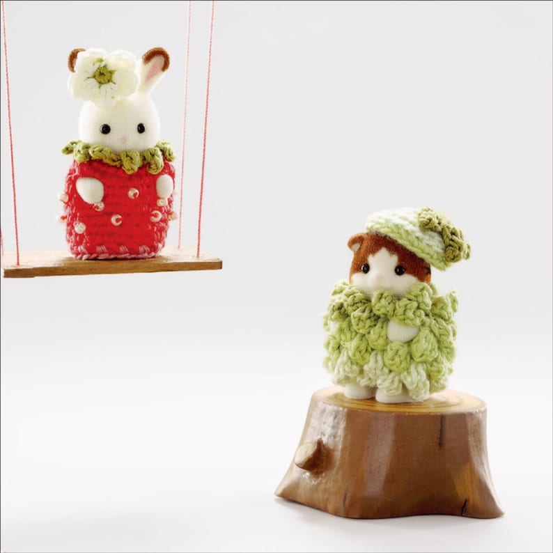 Pode incluir: Uma figura de coelho branco vestindo uma roupa de morango de croch&ecirc; vermelha com uma flor branca na cabe&ccedil;a, sentada em um balan&ccedil;o de madeira. Ao lado, uma figura de gato marrom e branco usa uma roupa e chap&eacute;u de croch&ecirc; verde claro sobre um toco.