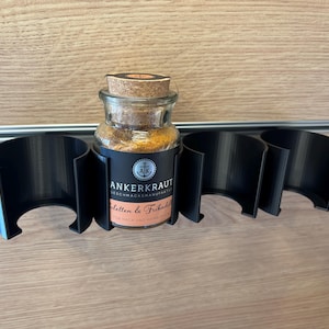 Könnte beinhalten: Ein klares Gewürzglas mit Korkdeckel, gefüllt mit gelben Gewürzen, steht in einem schwarzen Halter. Das Glas hat ein Etikett mit Text. Der Halter hat vier Abschnitte. Der Hintergrund ist hellbraunes Holz.