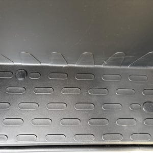 Könnte beinhalten: Eine schwarze Automatte mit einer strukturierten Oberfläche und einem sich wiederholenden Muster aus horizontalen ovalen Vertiefungen. Die Oberkante der Matte weist eine Reihe von sechs markanten, erhabenen dreieckigen Laschen auf.