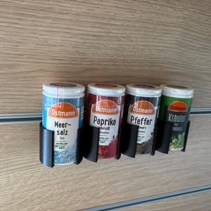 Könnte beinhalten: Vier Gewürzbehälter in schwarzen Haltern sind auf einer Holzoberfläche montiert. Die Behälter sind mit deutschem Text beschriftet, darunter "Meer-salz", "Paprika", "Pfeffer" und "Kräuter". Die Behälter haben weiße Deckel und farbige Etiketten.