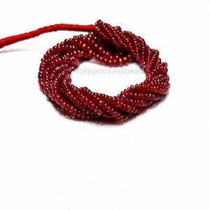 May include: A circular arrangement of small, deep red, faceted gemstone beads strung together on a thin red cord. The beads are uniform in size and shape, creating a dense, textured ring of dark crimson color against a plain white background.