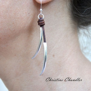 May include: Silver and brown leather dangle earrings. The earrings feature a long, curved silver piece with a brown leather cord wrapped around the top.