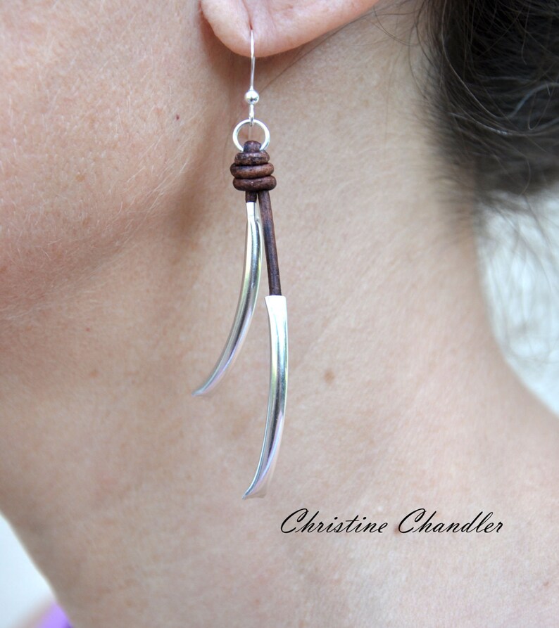 May include: Silver and brown leather dangle earrings. The earrings feature a long, curved silver piece with a brown leather cord wrapped around the top.