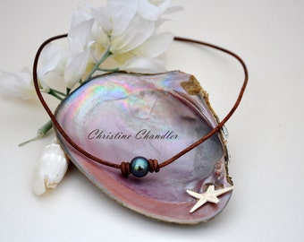 Puede incluir: Un collar de cordón de cuero marrón con una perla azul oscuro y un dije de estrella de mar plateada. El collar se muestra en una concha de nácar.