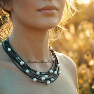 May include: A multi-strand necklace featuring black cords and luminous, round, white pearls. The necklace is layered and rests on a person's neck. The background is a blurred, sunlit field. The necklace is signed "Christine Chandler".