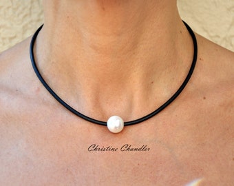 Puede incluir: Un collar de gargantilla negro con un solo colgante de perla blanca grande. El collar se lleva alrededor del cuello de una persona. El texto "Christine Chandler" está escrito en cursiva debajo.