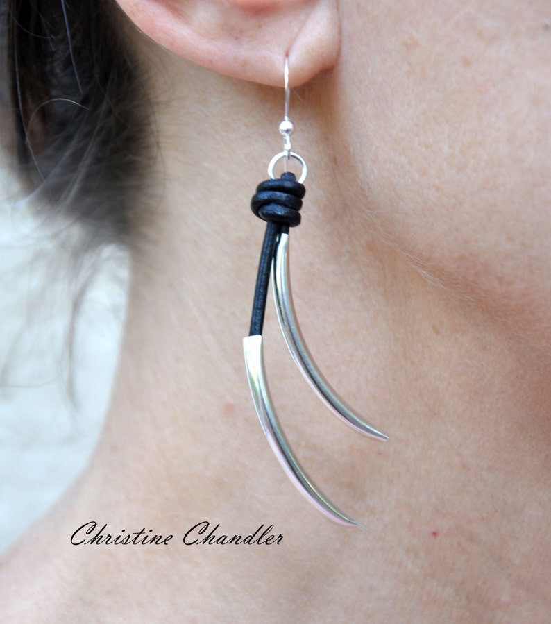 May include: Silver earrings with a black leather cord and a knot at the top. The earrings are long and have a curved shape.